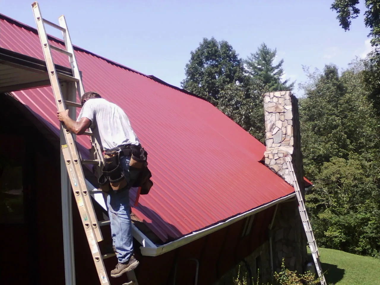 Roofer on a ladder beside a red metal roof and stone chimney — Soffit & Fascia Repair by Roofing Brothers in North Lawrence